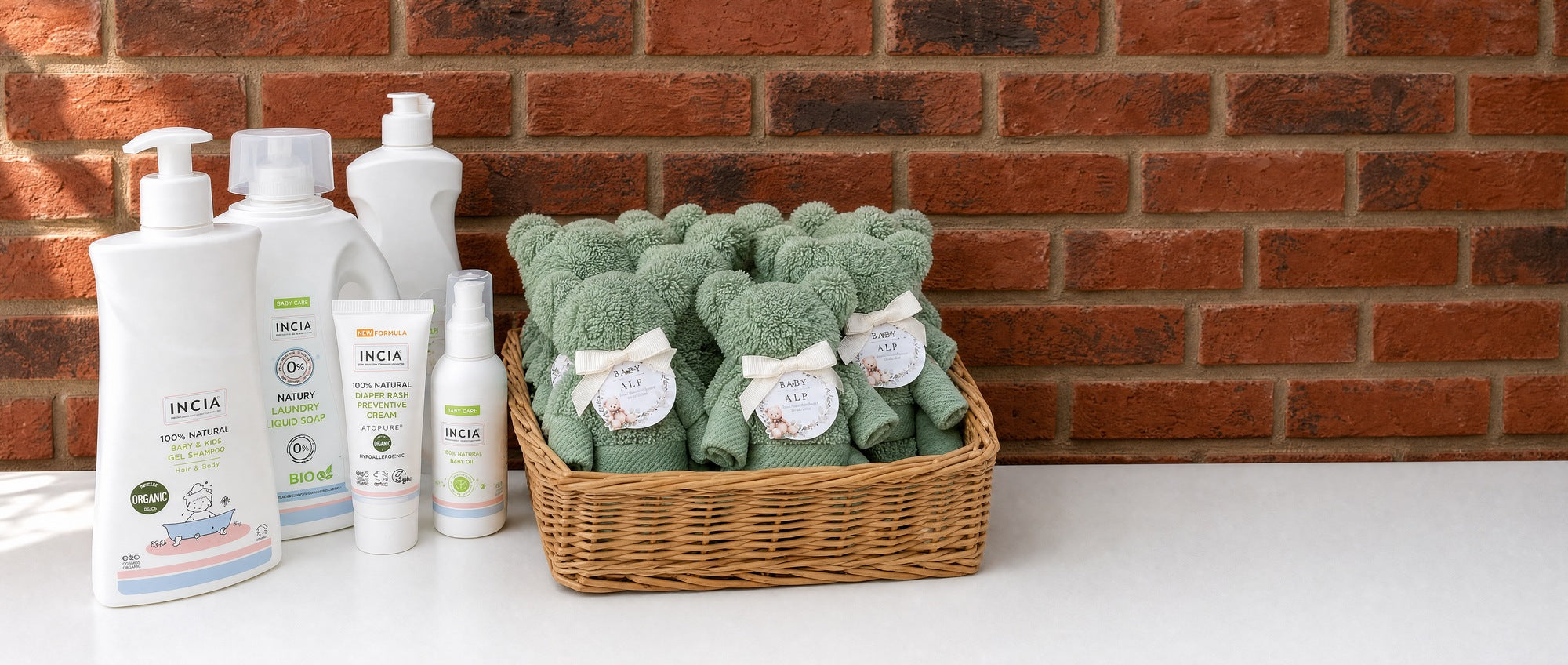 INCIA baby care bottles beside a basket of personalised sage-green towel bears against a brick wall